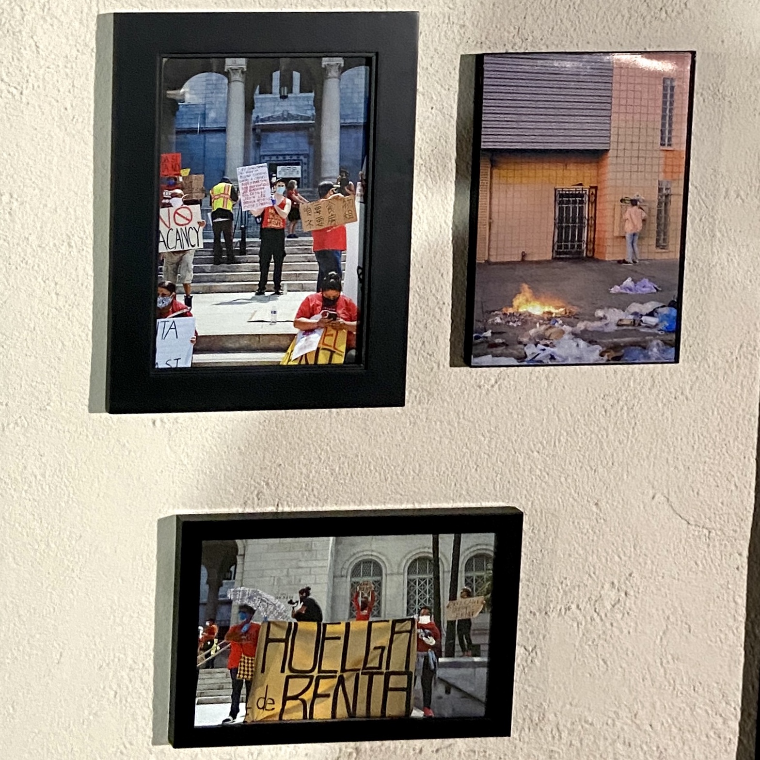 Three framed photos on a wall. Two are of people holding signs in front of City Hall with messages like 'No Vacancy' and 'Huelga de Renta.'